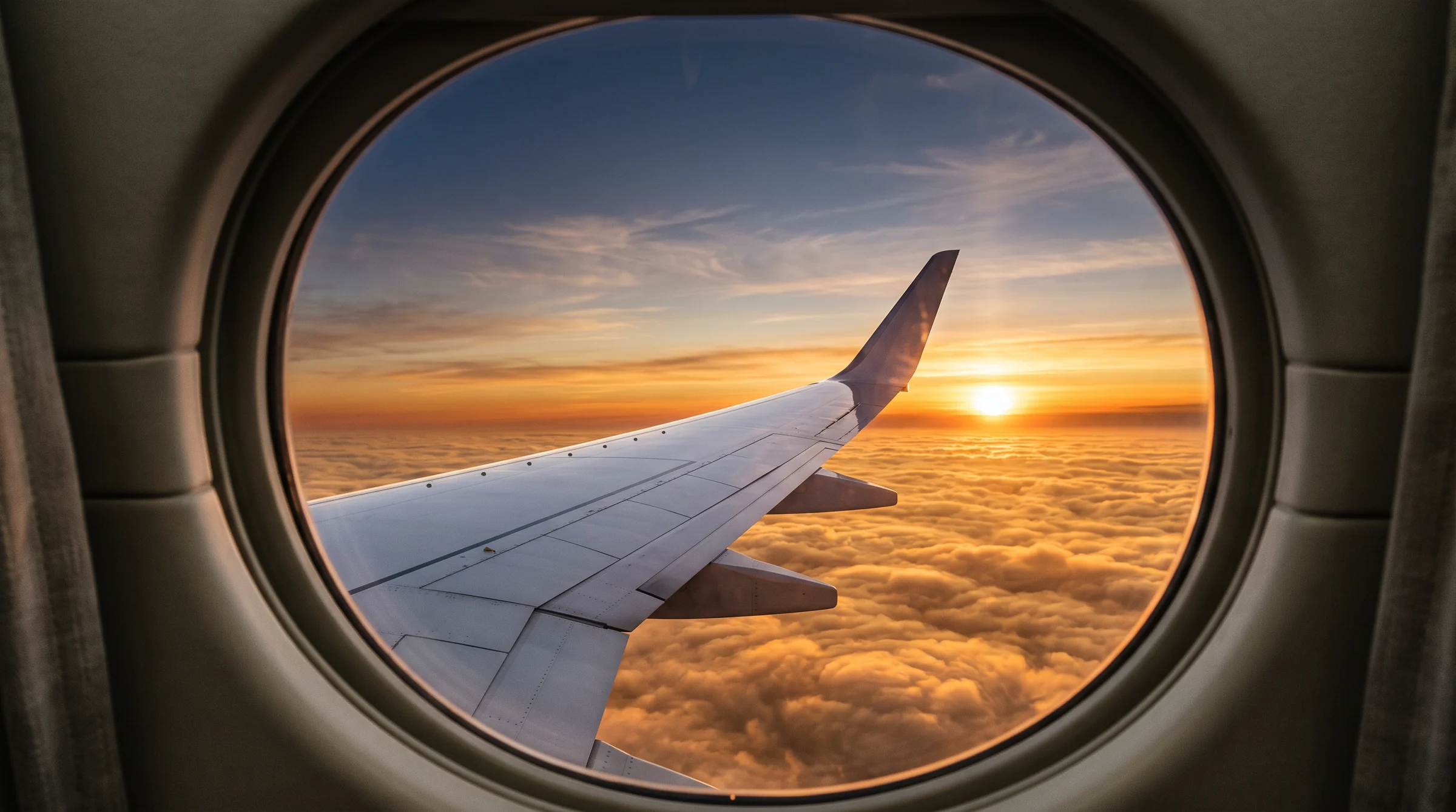 A wing and winglet seen through an oval cabin window above golden clouds